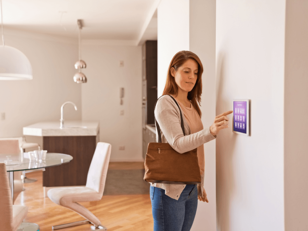 A woman stands in a modern living room, wearing a brown bag and touching a wall-mounted smart home control panel—showcasing vacation home security for safer summer travel.