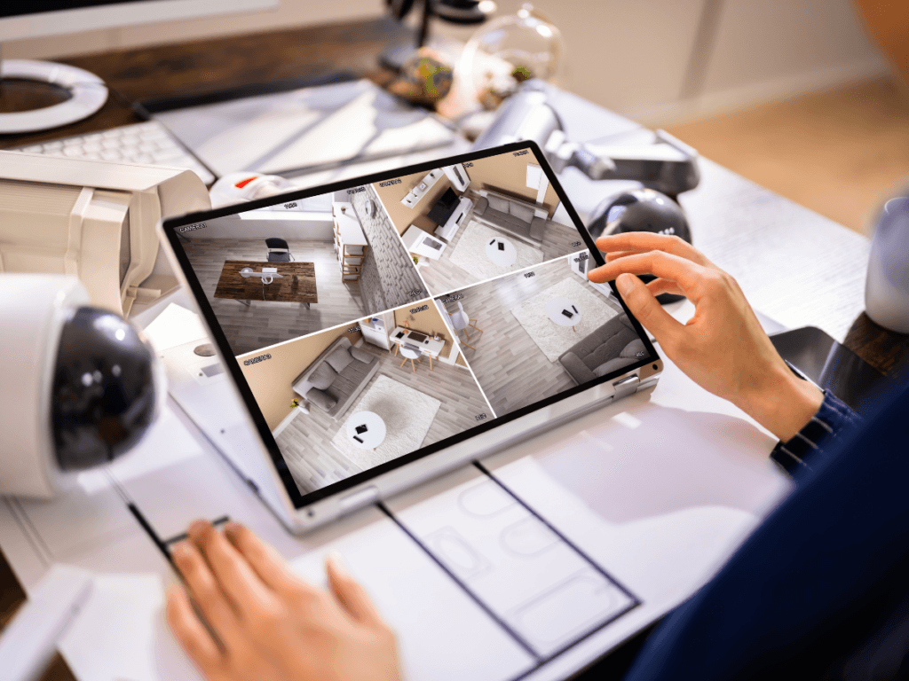 Person monitoring four indoor security camera feeds on a tablet at a desk with surveillance equipment and paperwork, demonstrating effective Vacation Home Security and Security System Tips.