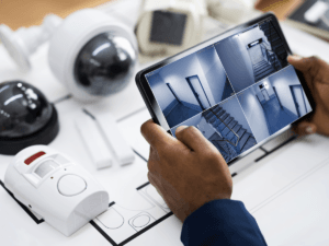 Person holding a tablet displaying live security camera footage of hallways and staircases, with various surveillance cameras and blueprints visible on the desk—perfect for checking vacation home security during summer travel.