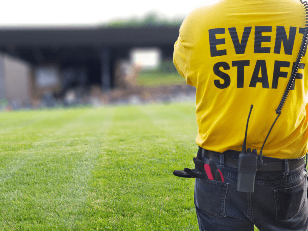 A concert security staff member in a yellow shirt stands on grass facing a blurred outdoor stage, wearing a radio and carrying tools on their belt.