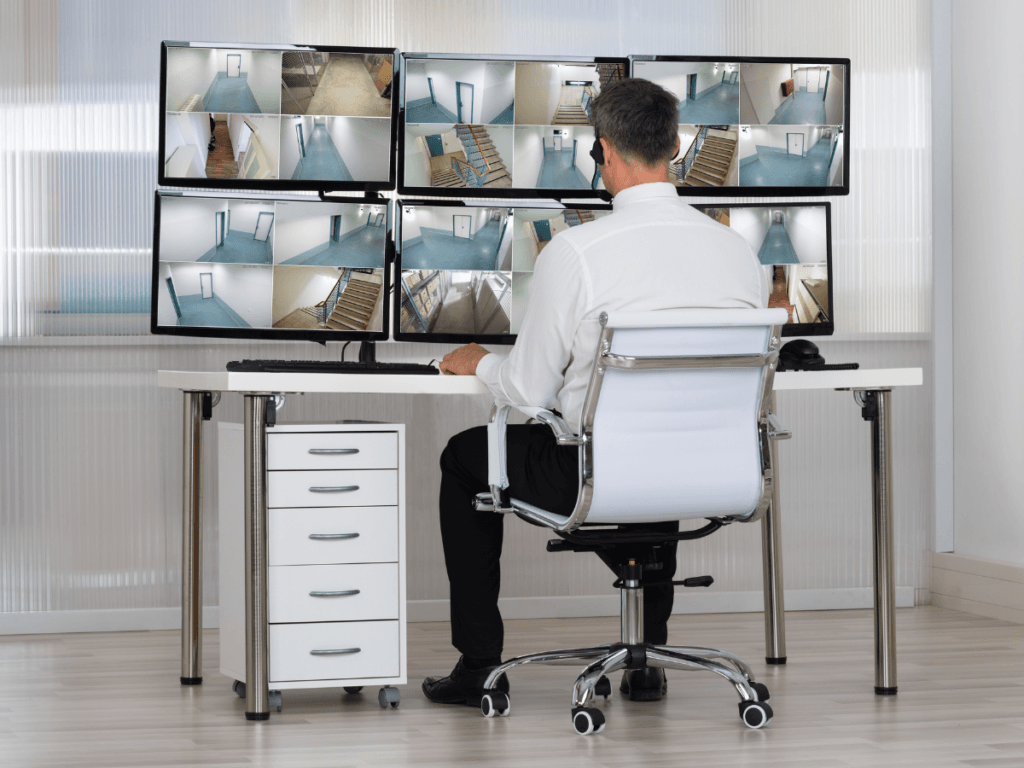 A man sits at a desk monitoring multiple surveillance camera feeds displayed on two rows of screens in a security control room, showcasing professional Security Services in College Park.
