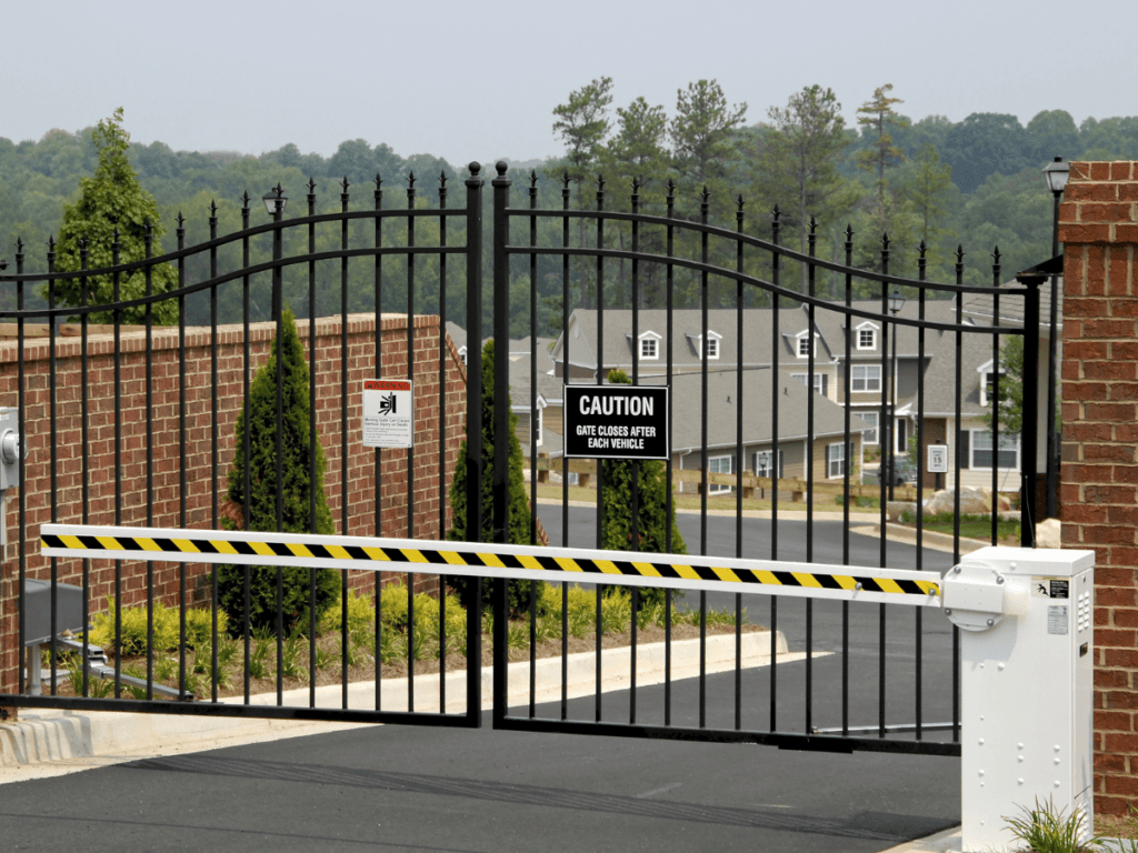 Automated security gate with warning signs and barrier arm at the entrance of a residential neighborhood, showcasing robust gated community security with houses and trees visible in the background.