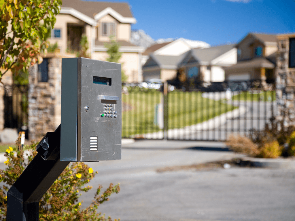 A keypad entry system stands in front of a closed security gate at the entrance to a gated community, ensuring enhanced security for the residential neighborhood with houses visible in the background.