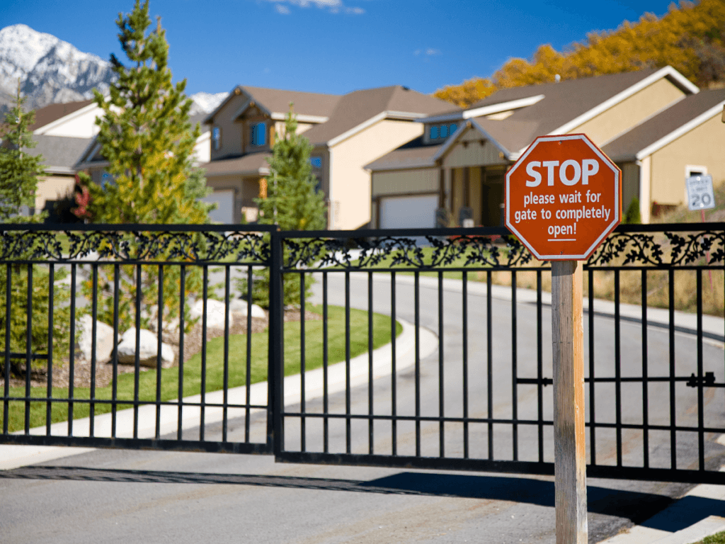 A closed metal gate—part of the gated community security measures—blocks the entrance to a residential neighborhood. A red sign reads, “STOP please wait for gate to completely open!” Houses and trees are visible in the background.