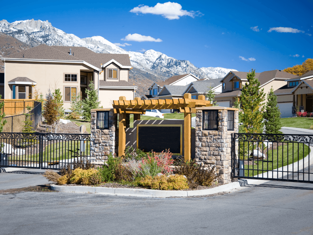 Gated entrance to a suburban neighborhood with stone pillars, wooden pergola, and landscaped plants, offering gated community security, with houses and snow-capped mountains in the background under a clear sky.