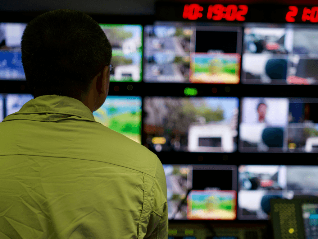 Person facing multiple monitors in a control room, displaying various video feeds from comprehensive surveillance services, with digital clocks visible at the top of the screens.