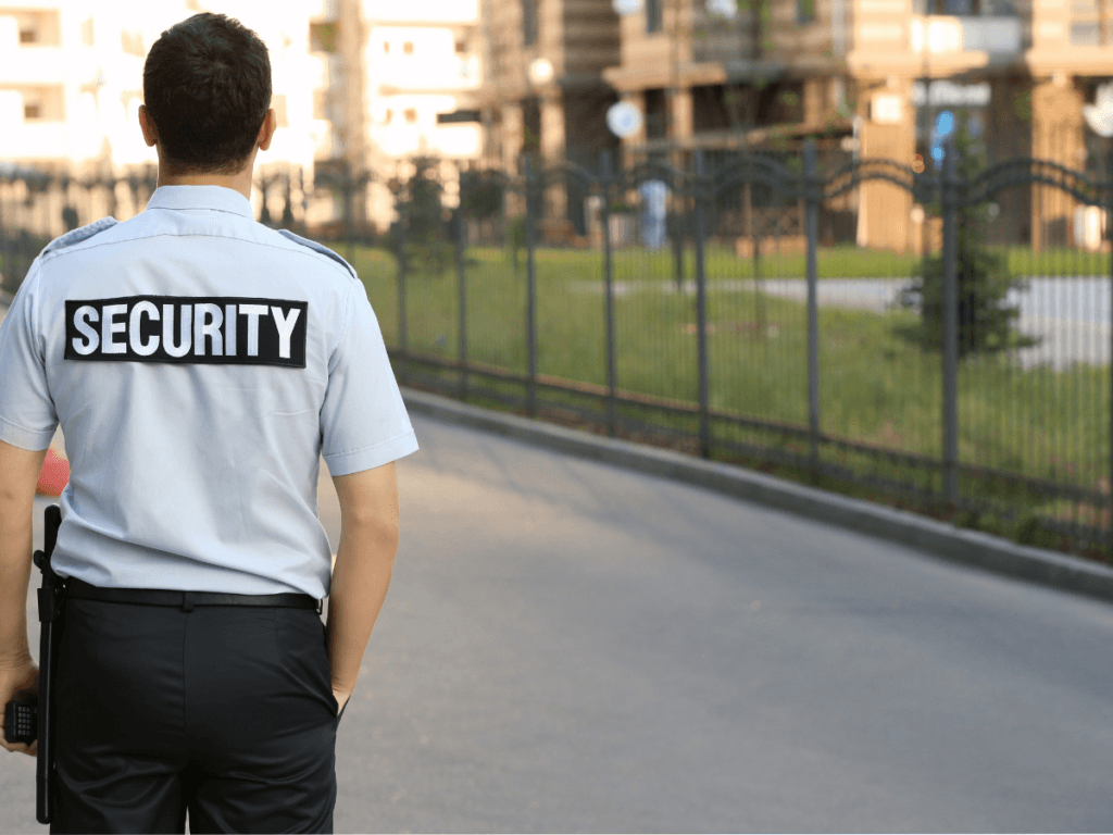 A campus security guard in uniform stands outside on a paved path near a fenced area, facing away from the camera.