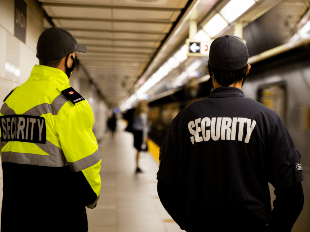Two campus security guards stand on a subway platform, facing the tracks; one wears a yellow jacket, the other a dark jacket, both labeled “SECURITY” on the back.