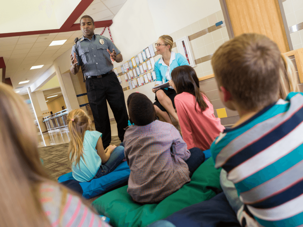 A campus security officer speaks to a group of children seated on the floor in a hallway, while an adult sits nearby taking notes.