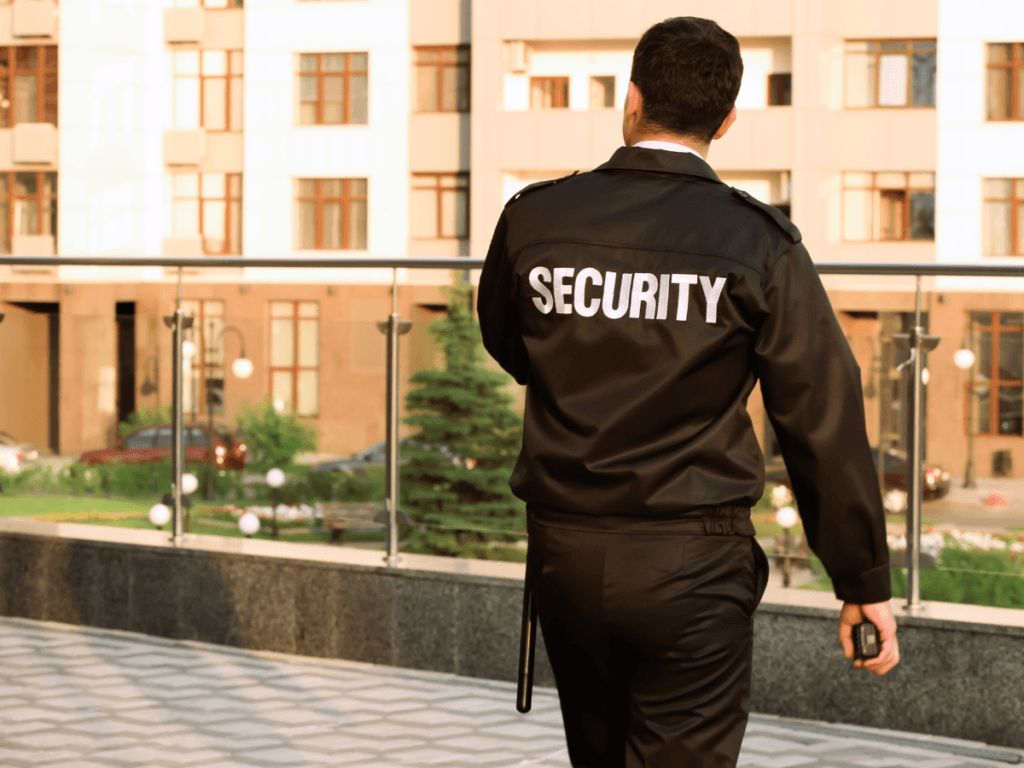 A campus security guard in uniform stands outdoors, holding a walkie-talkie, facing away from the camera with a building and greenery in the background.