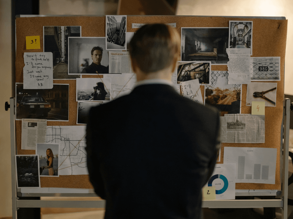 A person in a suit stands facing a corkboard covered with photos, notes, maps, and newspaper clippings, suggesting an intense domestic investigation.