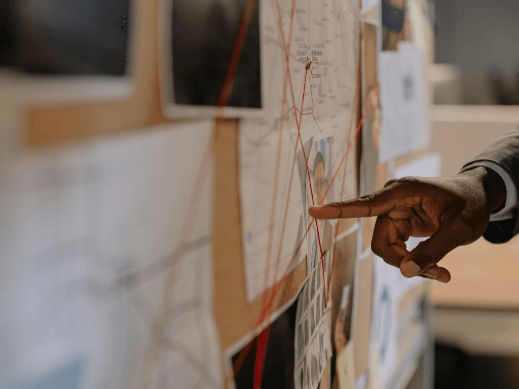 A person points at a board covered with maps, photos, and red string connecting various items, suggesting an investigation or analysis—possibly related to domestic investigations.